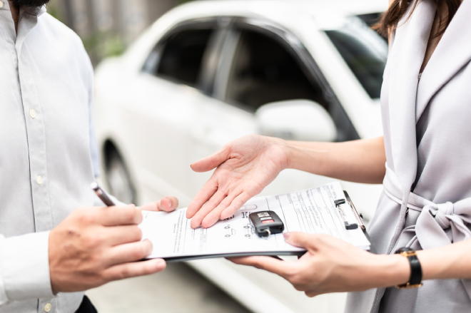 A man signs paperwork whilst buying a car, and a woman shows him where to sign.