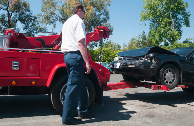 A man operates a red tow truck as it lifts a crashed black car