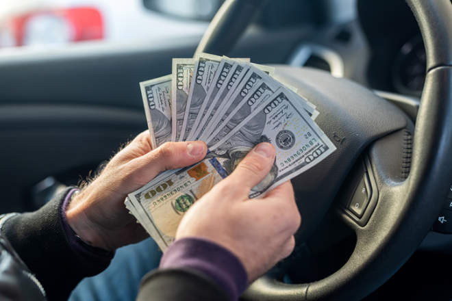 A man fans some US dollar bills in front of a car steering wheel